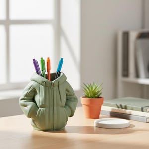 Op de afbeelding: Een saliegroene pennenhouder in de vorm van een jas met capuchon, met een rits en zakken. Het bevat een paarse, groene, oranje en blauwe pen. Een kleine potplant en een witte onderzetter staan ook op het houten bureau.