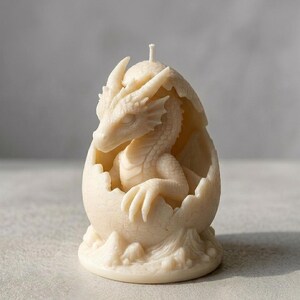 Könnte beinhalten: Eine cremefarbene Kerze in Form eines Drachen, der aus einem Ei schlüpft. Der Drache hat detaillierte Schuppen, Hörner und Krallen. Die Eierschale ist strukturiert, und oben befindet sich ein kleiner Docht. Diese dekorative Kerze ist für Fantasy-Enthusiasten.