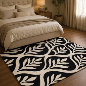 May include: A black and white patterned area rug with a leaf design. The rug is placed in a bedroom setting, with a bed and nightstand visible in the background. The rug's design features a repeating pattern of stylized leaves.