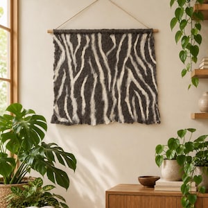 Könnte beinhalten: Ein quadratischer Wandbehang mit einem Zebramuster in Dunkelanthrazit und Cremeweiß. Das Textil ist an einem horizontalen Holzstab aufgehängt, an dessen Enden eine dünne Schnur zur Befestigung an einer Wand angebracht ist.