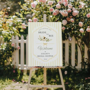 May include: A rectangular sign with vertical yellow and white stripes featuring the text Let's Honor The Bride To Bee, Welcome to Lillie's Bridal Shower, and the date 09.15.26, decorated with a small bee illustration and floral accents.