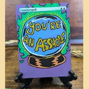 Könnte beinhalten: Eine rechteckige Sammelkarte mit einer handgemalten Illustration einer blauen Kristallkugel, die von grünen Flammen vor einem violetten Hintergrund umgeben ist. Der Text "Counterspell" steht oben und "YOU'RE AN ASSHOLE" ist in Gelb geschrieben.