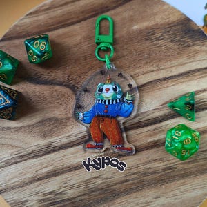 Puede incluir: Un llavero acrílico transparente con un personaje de payaso de dibujos animados con un cuello a rayas verdes y blancas, una chaqueta azul y pantalones naranjas. El llavero está sujeto a un cierre verde. Varios dados verdes con números dorados están esparcidos sobre una superficie de madera. La palabra "kypas" está escrita en cursiva.