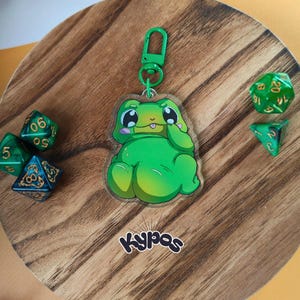 Könnte beinhalten: Ein grüner Acryl-Frosch-Schlüsselanhänger mit einem grünen Verschluss. Der Frosch hat große Augen und hält seine Hand vor sein Gesicht. Mehrere grüne Würfel sind um den Schlüsselanhänger auf einer Holzoberfläche verstreut. Das Wort "kypas" ist unten aufgedruckt.