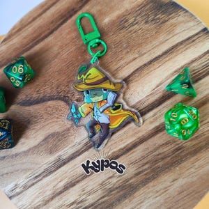 Può includere: Un portachiavi in acrilico trasparente con un cartone animato di una rana mago con cappello e cappotto gialli, una bacchetta blu e un fermaglio verde. Diversi dadi verdi e blu sono sparsi intorno al portachiavi su una superficie di legno. La parola "Kypas" è stampata.