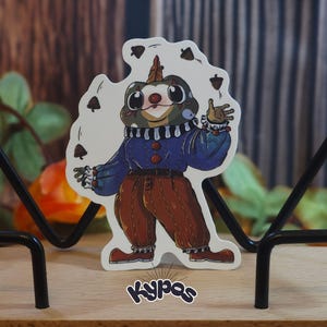 Op de afbeelding: Een sticker met een cartoonkikker verkleed als clown, die met eikels jongleert. De kikker draagt een blauwe jas, een bruine broek en rode schoenen. De sticker heeft onderaan de tekst "Kypas".