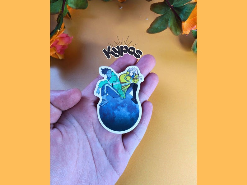 May include: A sticker featuring two anime-style figures sitting on a blue planet. One figure wears a green jacket, the other a yellow jacket. The sticker has the text "Kypas" in a stylized font.
