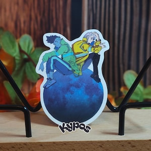 May include: A sticker featuring two anime-style characters sitting on a blue sphere. One character wears a green jacket and the other a yellow jacket. The sticker is displayed on a wooden base with a black metal stand. The sticker has the word "Kypas" on it.