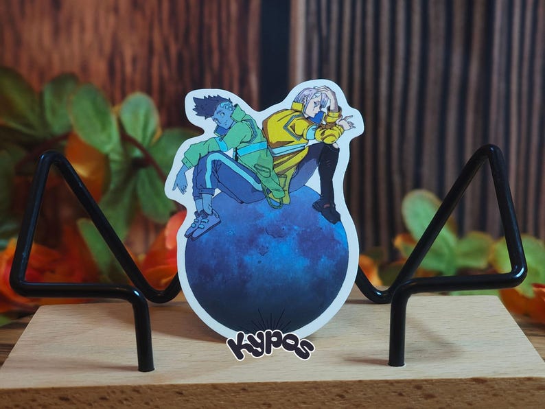 May include: A sticker featuring two anime-style characters sitting on a blue sphere. One character wears a green jacket and the other a yellow jacket. The sticker is displayed on a wooden base with a black metal stand. The sticker has the word "Kypas" on it.