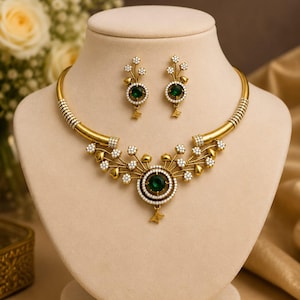 Peut inclure: Un ensemble collier et boucles d'oreilles doré composé de pendentifs circulaires avec des pierres vert foncé centrales, entourées de petites perles blanches et de branches métalliques dorées en forme de fleurs.