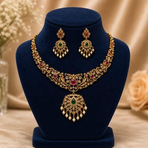 Può includere: Un set di collana e orecchini color oro con un intricato lavoro in filigrana metallica, incastonato con pietre rosse rotonde, un ciondolo centrale a goccia di colore verde e piccole perline bianche pendenti dai bordi dei gioielli.