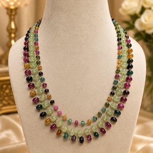 May include: A three-strand necklace featuring small, polished, multi-coloured gemstone beads in shades of light green, deep pink, dark blue, and golden yellow. The beads are strung on a thin gold-toned wire, creating a layered, circular design.