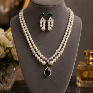 Puede incluir: Un collar de perlas de doble hebra con un colgante central en forma de lágrima de color verde esmeralda y pendientes a juego. El conjunto de joyería incluye pequeñas piedras transparentes engastadas en metal plateado, presentadas juntas.