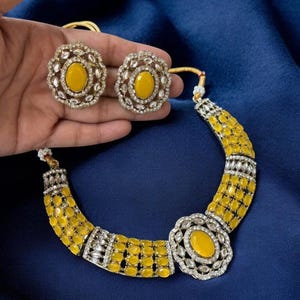 May include: A jewelry set consisting of a necklace and two stud earrings. The necklace features a central oval yellow stone surrounded by clear crystals, flanked by rows of yellow oval stones and clear crystal accents. Matching earrings included.