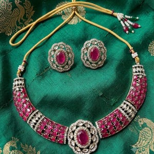 May include: A necklace and matching pair of stud earrings featuring oval-shaped deep pink stones set in silver-toned metal with clear accent stones. The necklace has a central oval pendant and a gold-toned adjustable cord with beaded tassels.