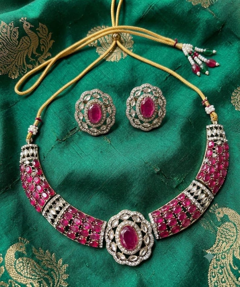 May include: A necklace and matching pair of stud earrings featuring oval-shaped deep pink stones set in silver-toned metal with clear accent stones. The necklace has a central oval pendant and a gold-toned adjustable cord with beaded tassels.