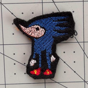 Op de afbeelding: Geborduurde patch met een blauw wezen met een lange snavel, een wit oog en rode voeten. De patch heeft een zwarte rand en staat op een grijze rasterachtergrond. De patch is ongeveer 3,8 cm hoog.
