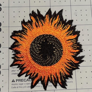 May include: Embroidered sunflower patch with a black center and orange petals. The patch is on a grid background with text that reads "A PRECAU" and other text in English and Spanish.