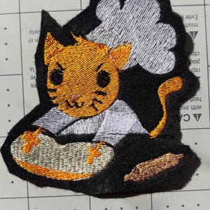 Può includere: Toppa ricamata raffigurante un gatto dei cartoni animati con pelo arancione, occhi neri e baffi. Il gatto indossa una camicia grigio chiaro ed è seduto su una superficie marrone chiaro. La toppa ha un bordo nero con un disegno di nuvola bianca.