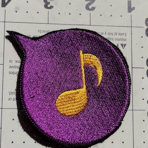 May include: A purple embroidered patch shaped like a speech bubble, with a gold musical note. The patch has a black border. It is set against a light grey surface with metric measurement markings.