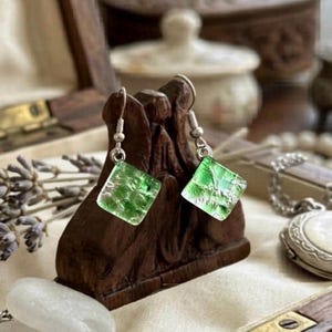 May include: A pair of square, green glass earrings with silver hooks. The earrings are displayed on a dark brown wooden sculpture. The earrings have a textured, translucent appearance, and are catching the light.