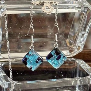 May include: A pair of square dangle earrings with a teal and dark purple speckled design. The earrings are suspended from silver-colored fish hook ear wires and chain. The earrings are displayed inside a clear, glass jewelry box.