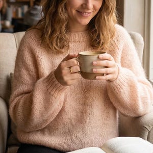 Puede incluir: Un suéter de punto rosa claro de gran tamaño con mangas largas. La persona sostiene una taza de cerámica beige con una base marrón. Se ve un anillo de oro en el dedo de la persona. La persona está sentada en un sillón beige, sosteniendo una taza de café.