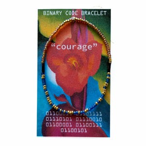 May include: A circular beaded bracelet featuring a repeating pattern of small metallic gold and blue beads. The bracelet is displayed on a rectangular card with the text BINARY CODE BRACELET, the word courage, and binary code sequences printed on it.