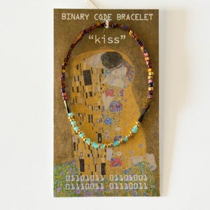 May include: A beaded bracelet featuring small metallic brown, gold, and turquoise beads arranged in a pattern. The bracelet is displayed on a rectangular card printed with the text "BINARY CODE BRACELET", "kiss", and binary sequences.