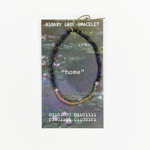 May include: A beaded bracelet displayed on a card labeled BINARY CODE BRACELET with the word home and binary sequences. The bracelet features small square and round beads in a gradient of dark blue, light blue, green, pink, and metallic gold.