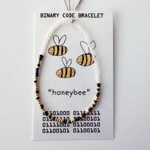 May include: A beaded bracelet featuring white, black, and gold beads arranged in a pattern on a white display card. The card displays the text "BINARY CODE BRACELET", "honeybee", and binary sequences, alongside three illustrated yellow and black bees.