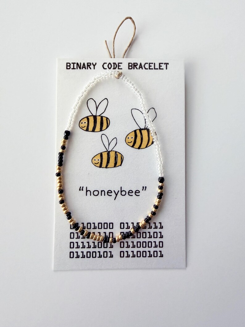 May include: A beaded bracelet featuring white, black, and gold beads arranged in a pattern on a white display card. The card displays the text "BINARY CODE BRACELET", "honeybee", and binary sequences, alongside three illustrated yellow and black bees.