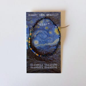 May include: A beaded bracelet featuring small dark blue, green, and gold beads arranged in a circular pattern on a display card. The card displays the text BINARY CODE BRACELET, "star", and binary digits, set against a starry night art print.