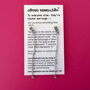 K&ouml;nnte beinhalten: Silberfarbene Ohrringe mit Kettenakzenten, pr&auml;sentiert auf einer wei&szlig;en Karte vor pinkfarbenem Hintergrund. Die Karte zeigt den Text "CODED REBELLION" und Produktinformationen. Die Ohrringe haben kleine herzf&ouml;rmige Anh&auml;nger.