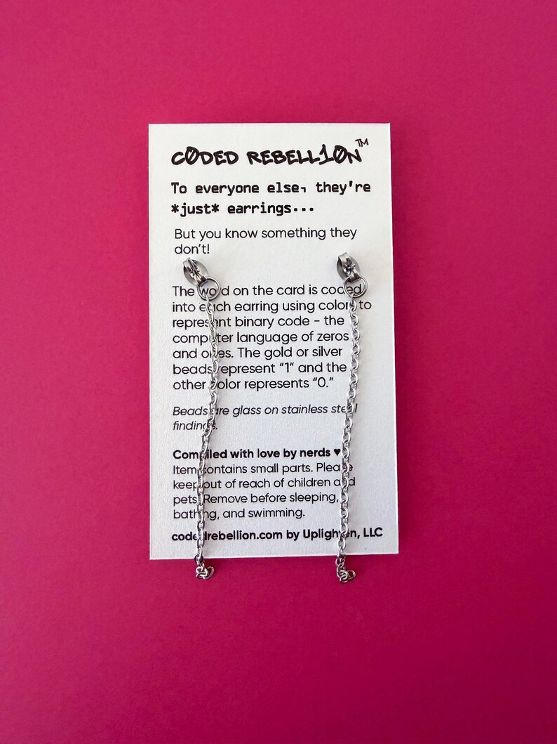 K&ouml;nnte beinhalten: Silberfarbene Ohrringe mit Kettenakzenten, pr&auml;sentiert auf einer wei&szlig;en Karte vor pinkfarbenem Hintergrund. Die Karte zeigt den Text "CODED REBELLION" und Produktinformationen. Die Ohrringe haben kleine herzf&ouml;rmige Anh&auml;nger.