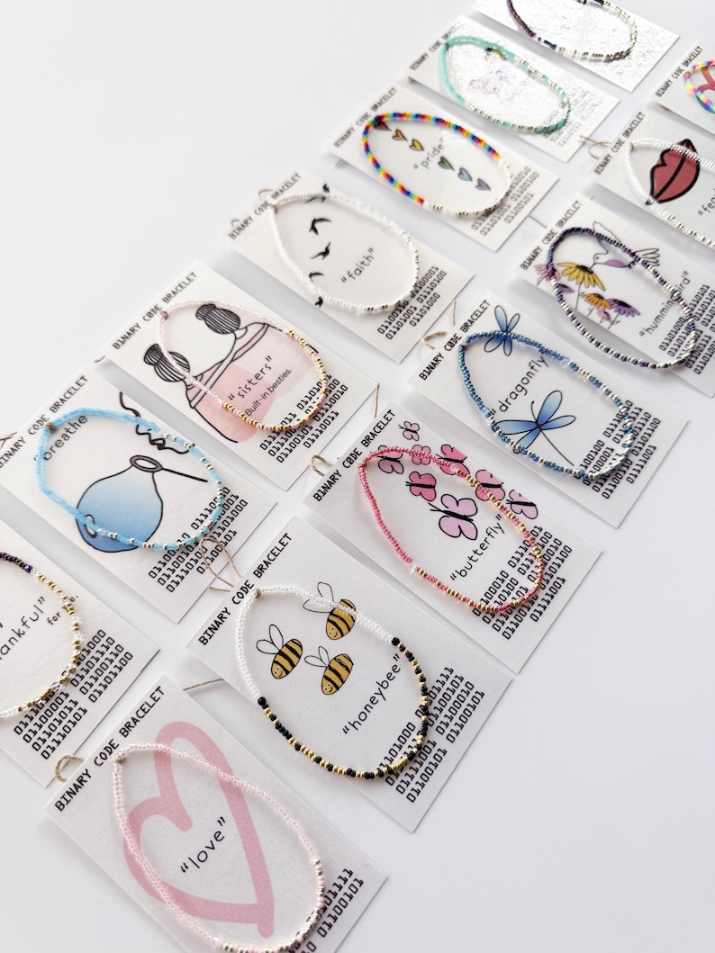 May include: A collection of beaded bracelets displayed on individual white cards, each featuring a unique graphic illustration and corresponding binary code text. The bracelets vary in color, including rainbow, blue, pink, black, and white beads.