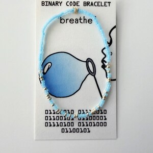 May include: A light blue and silver beaded bracelet displayed on a white card labeled BINARY CODE BRACELET. The card features a drawing of a person blowing a blue bubble and the word breathe, along with binary code text printed at the bottom.