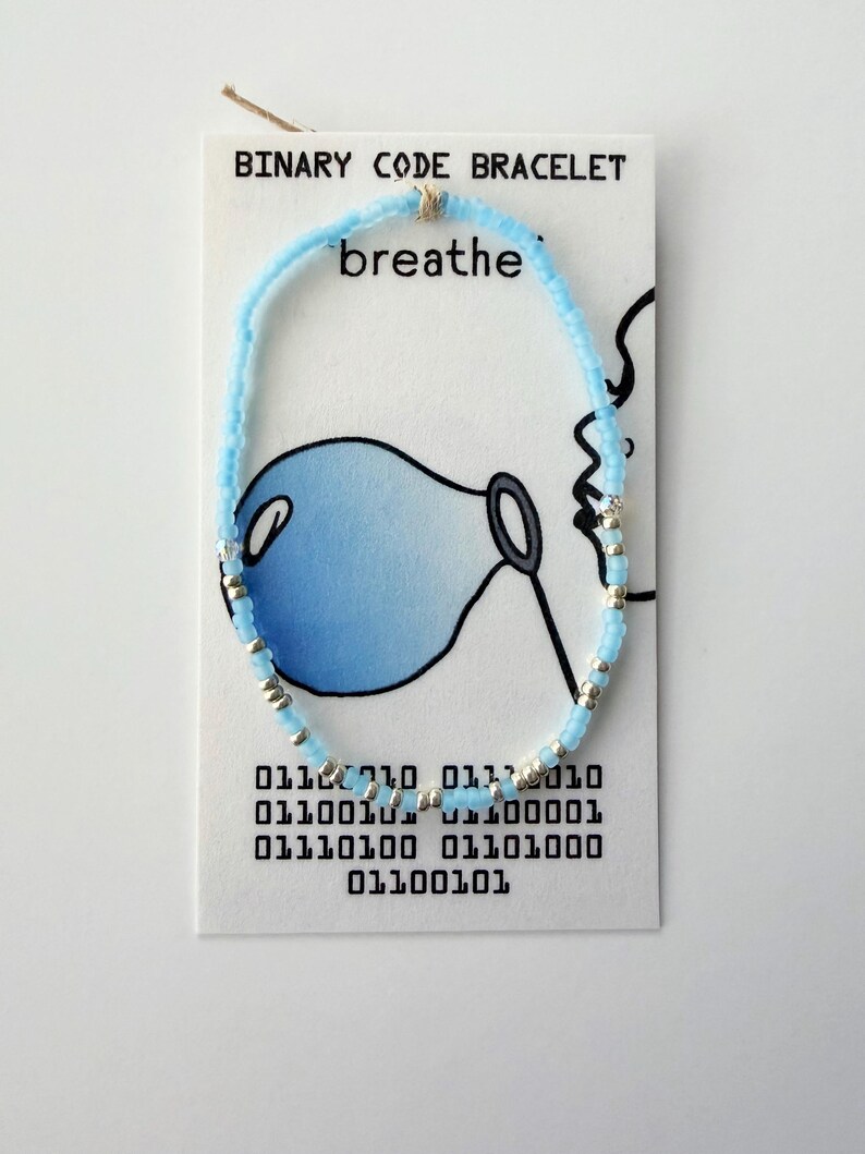 May include: A light blue and silver beaded bracelet displayed on a white card labeled BINARY CODE BRACELET. The card features a drawing of a person blowing a blue bubble and the word breathe, along with binary code text printed at the bottom.