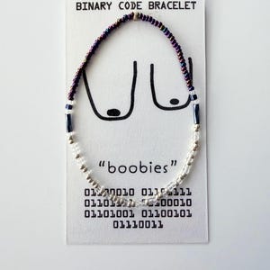 May include: A beaded bracelet on a white display card labeled BINARY CODE BRACELET. The bracelet features iridescent purple, dark blue, and clear beads. The card displays a line drawing of breasts, the word boobies, and binary code sequences.
