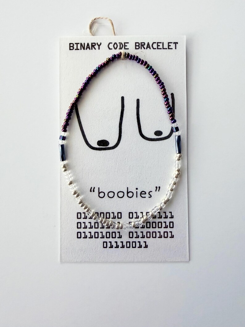 May include: A beaded bracelet on a white display card labeled BINARY CODE BRACELET. The bracelet features iridescent purple, dark blue, and clear beads. The card displays a line drawing of breasts, the word boobies, and binary code sequences.