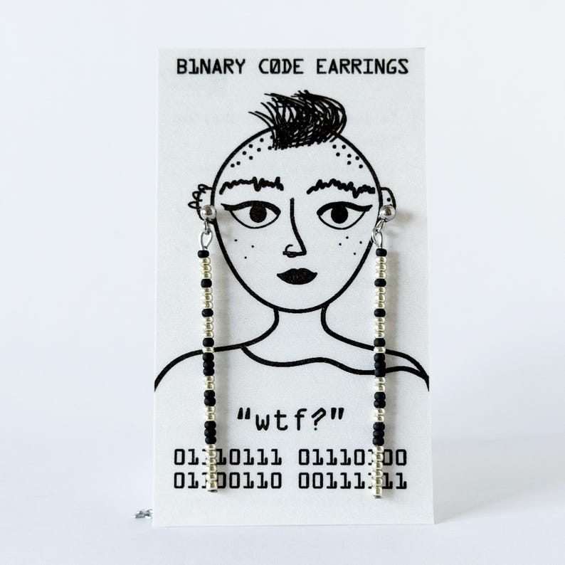 May include: A pair of long, linear earrings made of alternating black and silver beads, displayed on a white card featuring a black line drawing of a face, the text "BINARY CODE EARRINGS", "wtf?", and two lines of binary code digits.