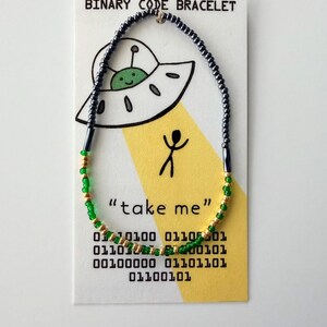 May include: A beaded bracelet featuring dark grey, green, and gold beads on a display card. The card shows a cartoon UFO, a stick figure, the text "BINARY CODE BRACELET" and "take me", and rows of binary code digits in black ink.