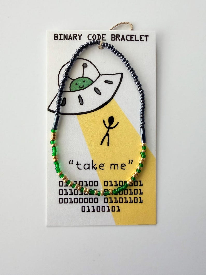 May include: A beaded bracelet featuring dark grey, green, and gold beads on a display card. The card shows a cartoon UFO, a stick figure, the text "BINARY CODE BRACELET" and "take me", and rows of binary code digits in black ink.