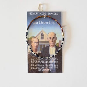 Könnte beinhalten: Ein kreisförmiges Perlenarmband mit einer Mischung aus metallischen, schwarzen und silbernen Perlen. Das Armband ist auf einer rechteckigen Karte mit der Aufschrift BINARY CODE BRACELET befestigt, die American Gothic und den Binärcode 01100001 01110101 01100101 01110100 01101001 01100110 01101001 01100001 01101100 zeigt.