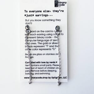 May include: Silver-colored chain earrings with a small charm at the end, displayed on a white card. The card has text that reads "To everyone else, they're *just* earrings..." and explains the earrings' binary code design.