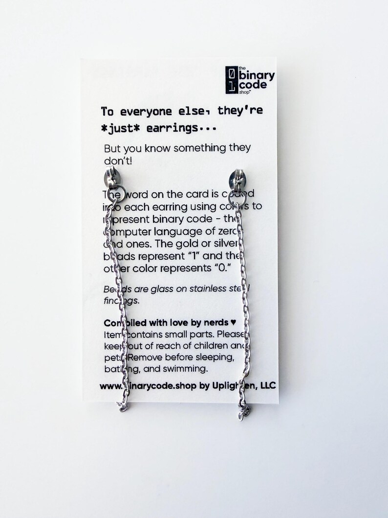 May include: Silver-colored chain earrings with a small charm at the end, displayed on a white card. The card has text that reads "To everyone else, they're *just* earrings..." and explains the earrings' binary code design.