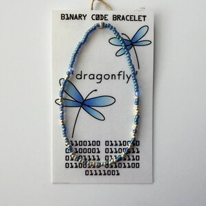 May include: A beaded bracelet featuring alternating blue and silver-toned beads on a white display card. The card displays the text BINARY CODE BRACELET, the word dragonfly, and binary code sequences, accompanied by illustrations of two blue dragonflies.