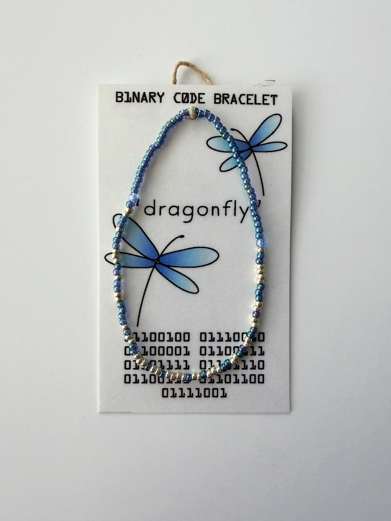 May include: A beaded bracelet featuring alternating blue and silver-toned beads on a white display card. The card displays the text BINARY CODE BRACELET, the word dragonfly, and binary code sequences, accompanied by illustrations of two blue dragonflies.