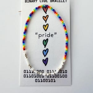 May include: A beaded bracelet featuring a rainbow-colored section and a white and silver section, displayed on a white card labeled BINARY CODE BRACELET. The card features five colorful hearts, the word pride, and binary code sequences.