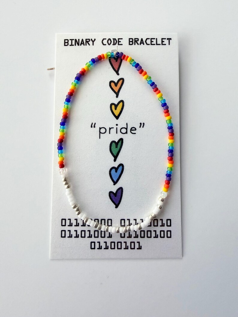 May include: A beaded bracelet featuring a rainbow-colored section and a white and silver section, displayed on a white card labeled BINARY CODE BRACELET. The card features five colorful hearts, the word pride, and binary code sequences.